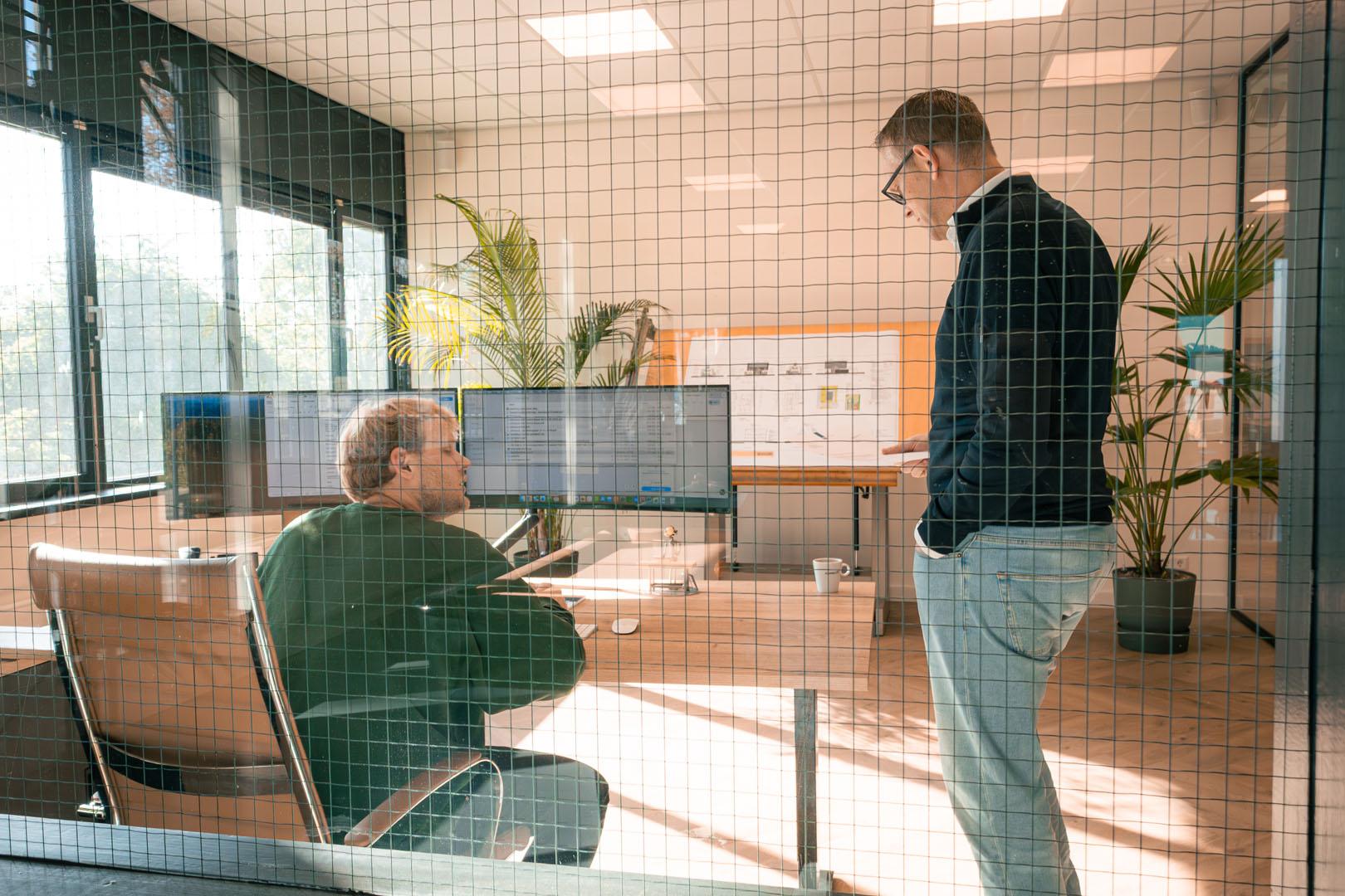 Twee personen bespreken werkvoorbereiding in kantoor met computerschermen