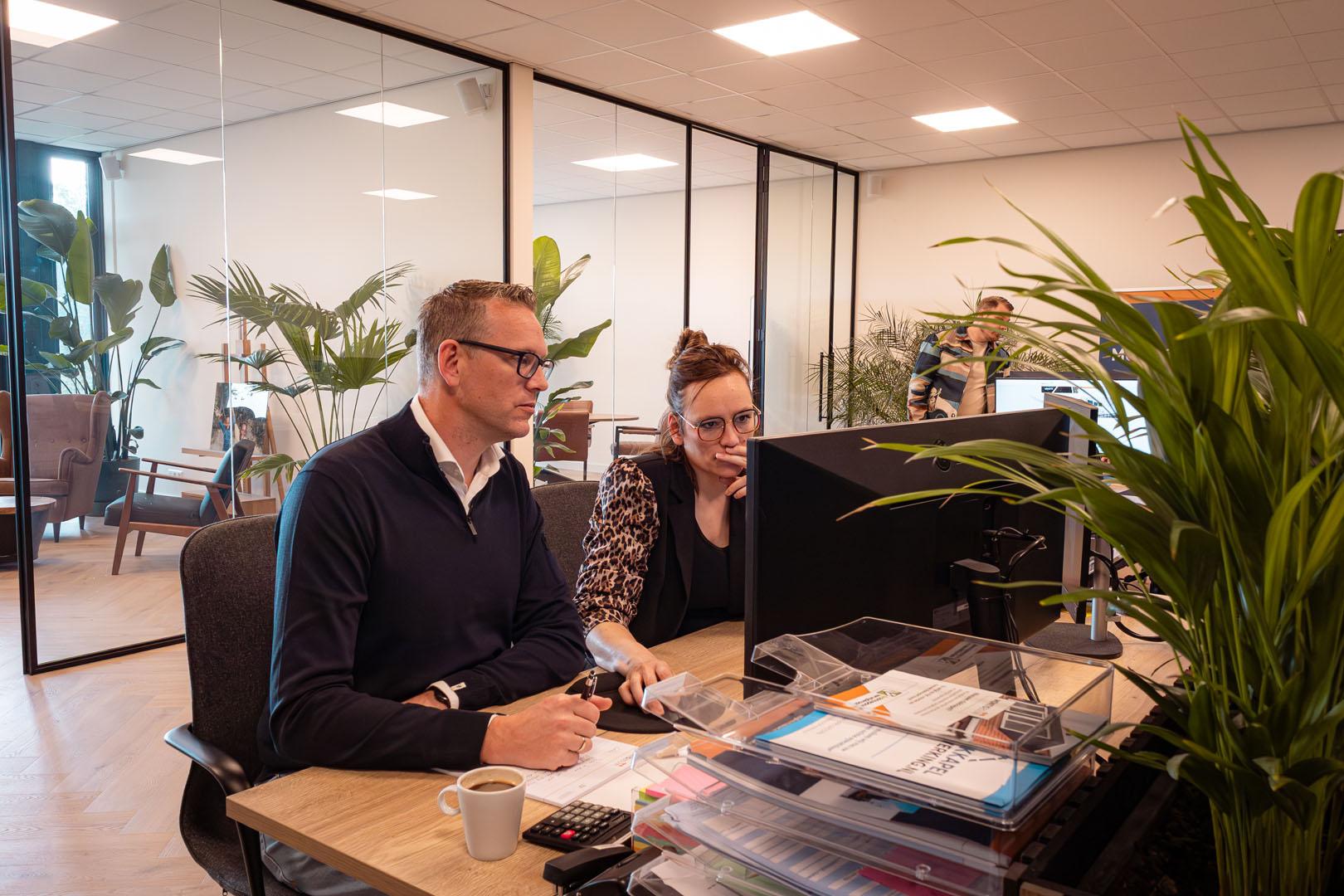 Twee werknemers werken samen aan een computermonitor met papieren en documenten op kantoor