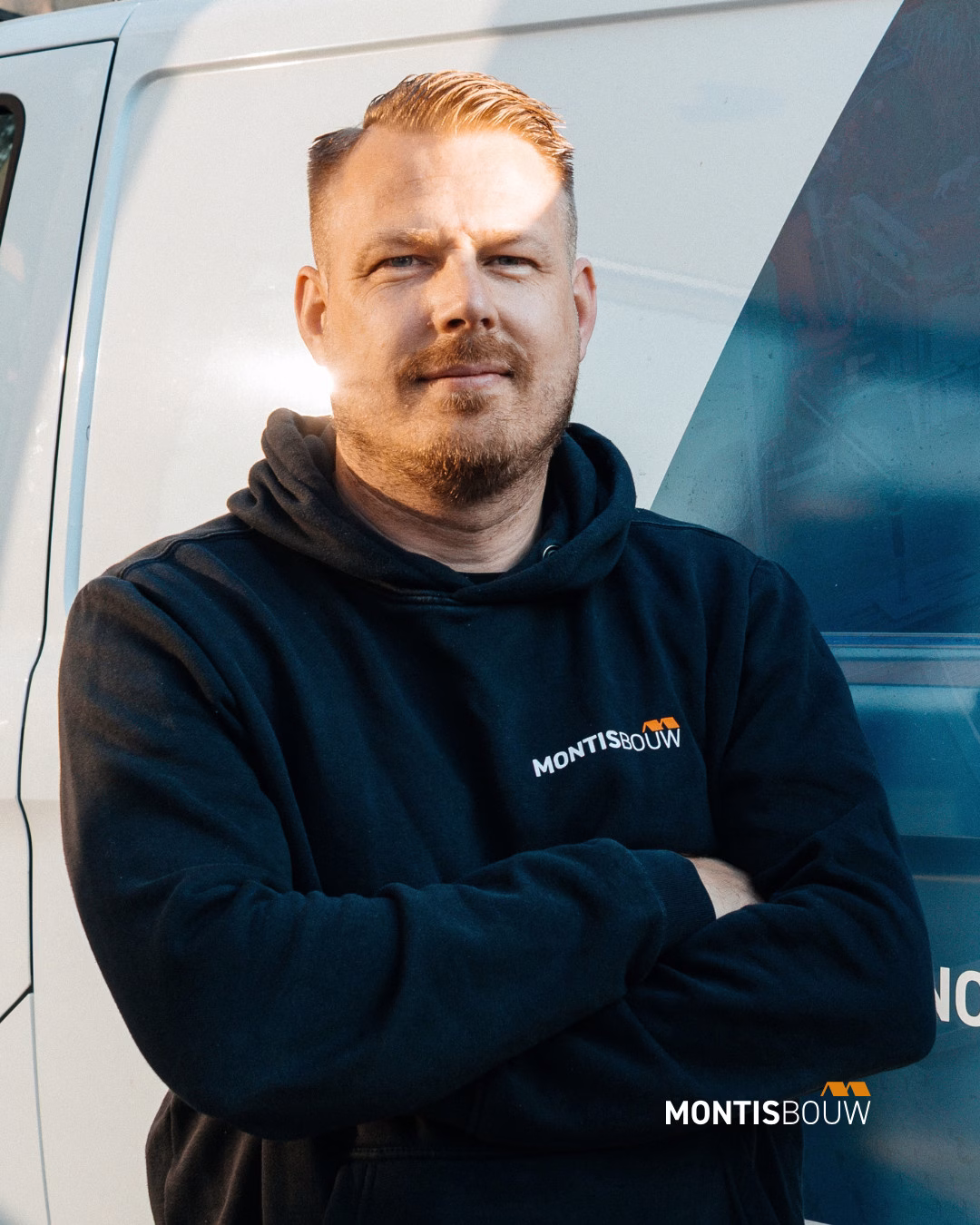 Man in MONTISBOUW-hoodie staat voor werkbus met armen gekruist