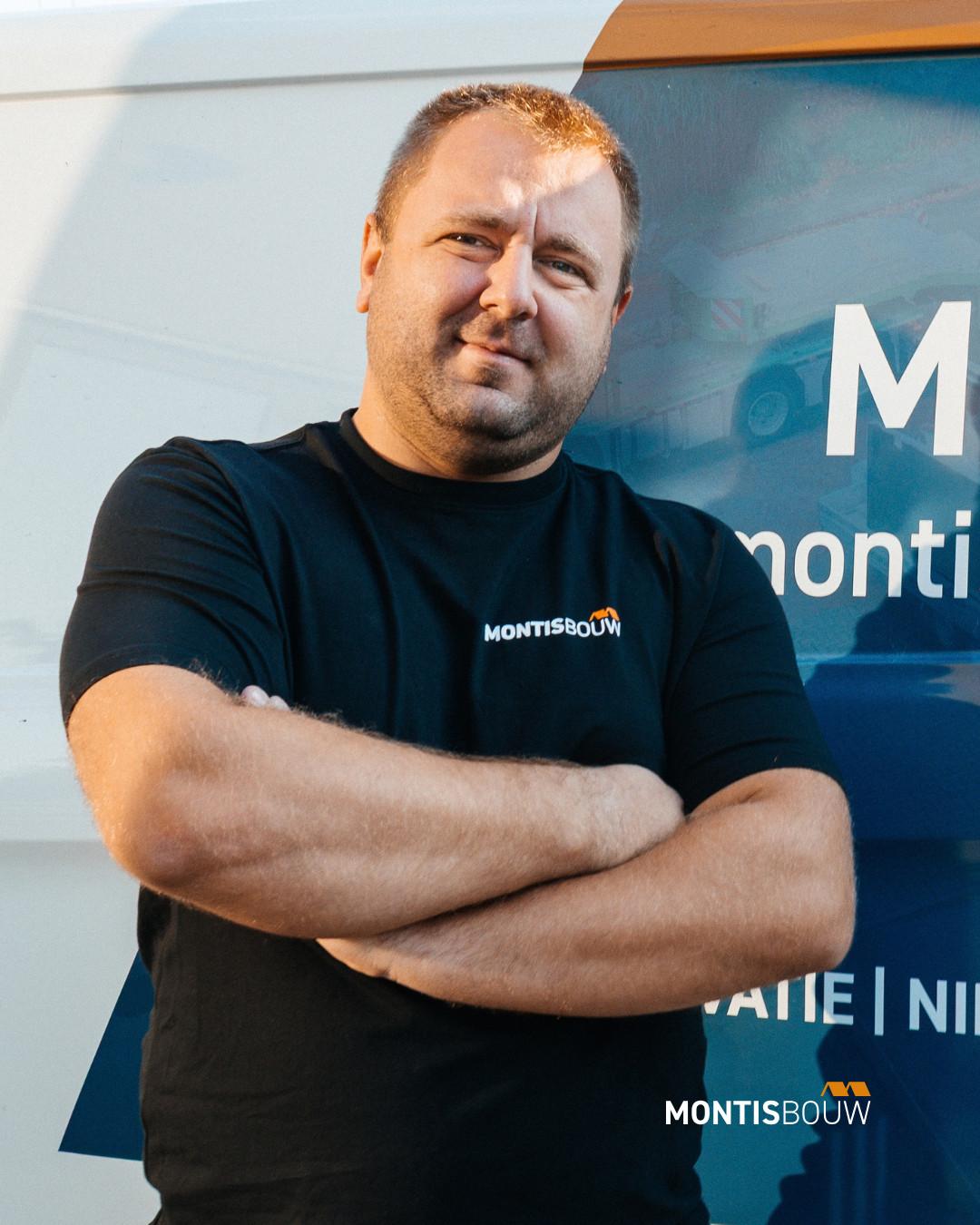 Man in zwart Montisbouw-shirt met gekruiste armen voor blauwe bedrijfsachtergrond